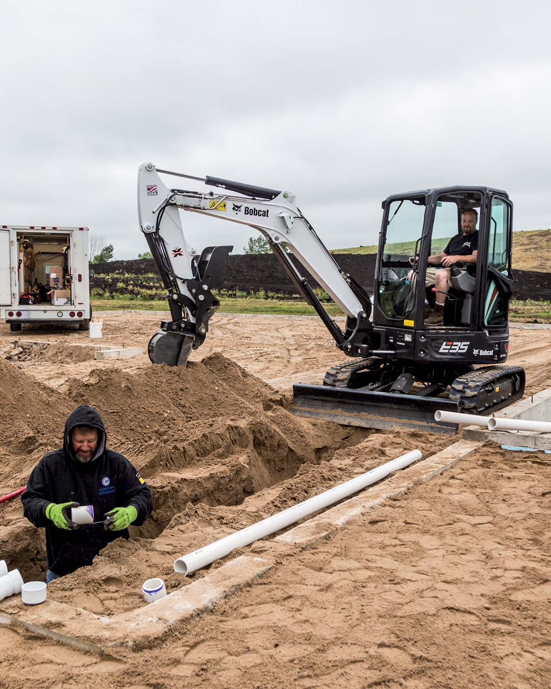 A Bobcat E35 mini excavator is seen at a worksite, with an operator in the cab using the machine to assist with a trenching project. A worker stands in the trench installing PVC pipes, while the excavator efficiently clears soil. A service truck is parked nearby, contributing to the construction activity.