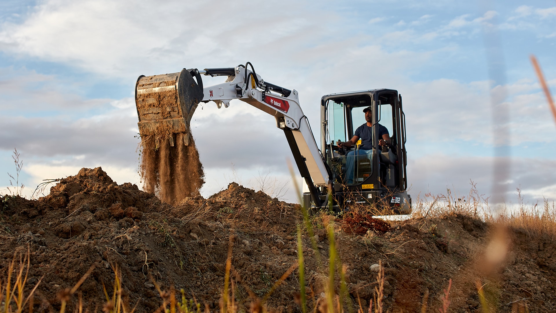 This image shows a Bobcat E32 mini excavator in action, a type of construction machinery ideal for tasks like digging, trenching, and material handling on construction sites. The excavator's operator is using the machine to scoop and dump dirt, showcasing its powerful hydraulic system and precise control. Its compact size allows for manoeuvrability in tight spaces while maintaining high productivity, making it a popular choice for small to mid-sized construction projects.