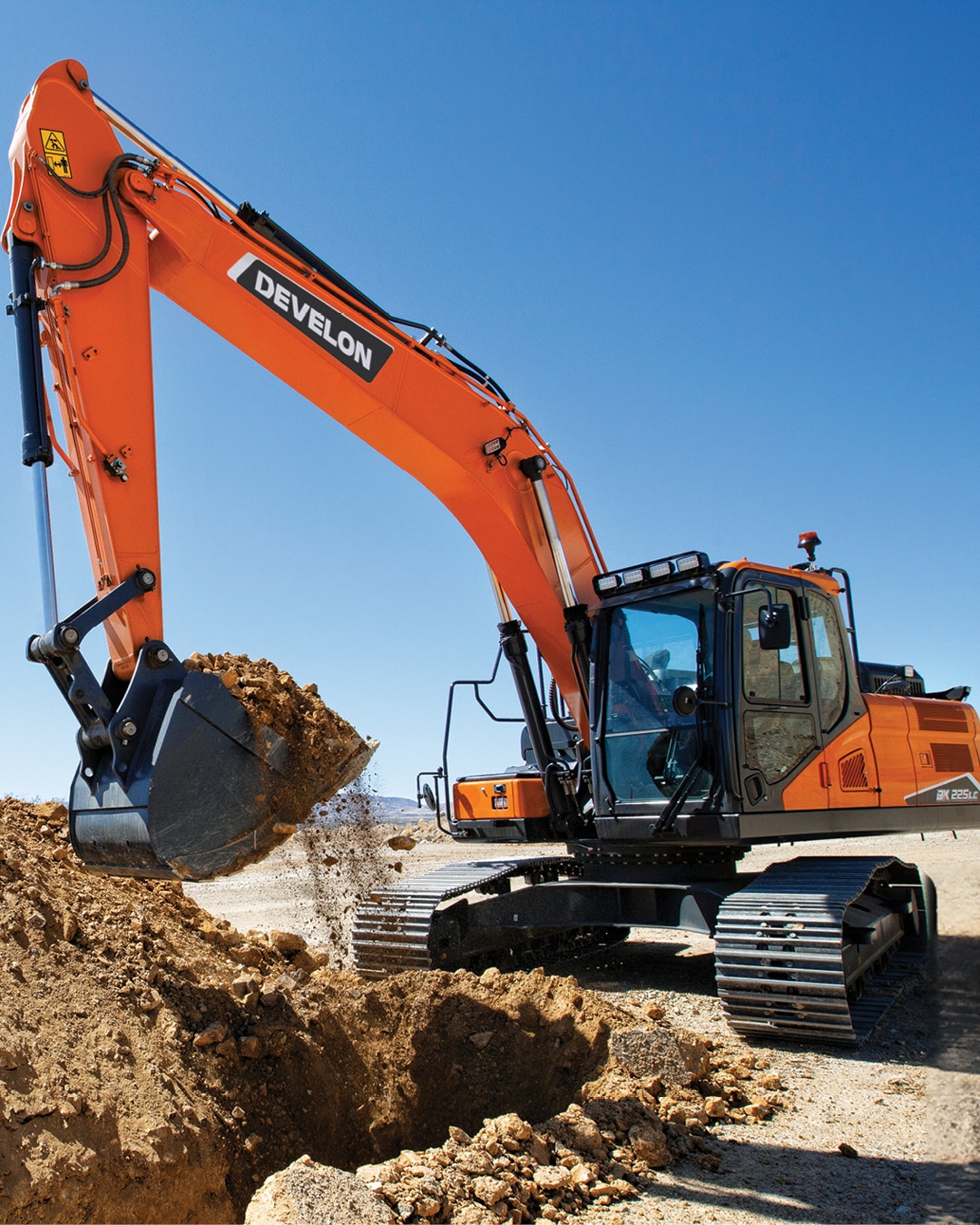 The image depicts a large earthmoving machinery unit, specifically a DEVELON-branded excavator (model DX225LC), actively scooping dirt from the ground with its bucket attachment.
