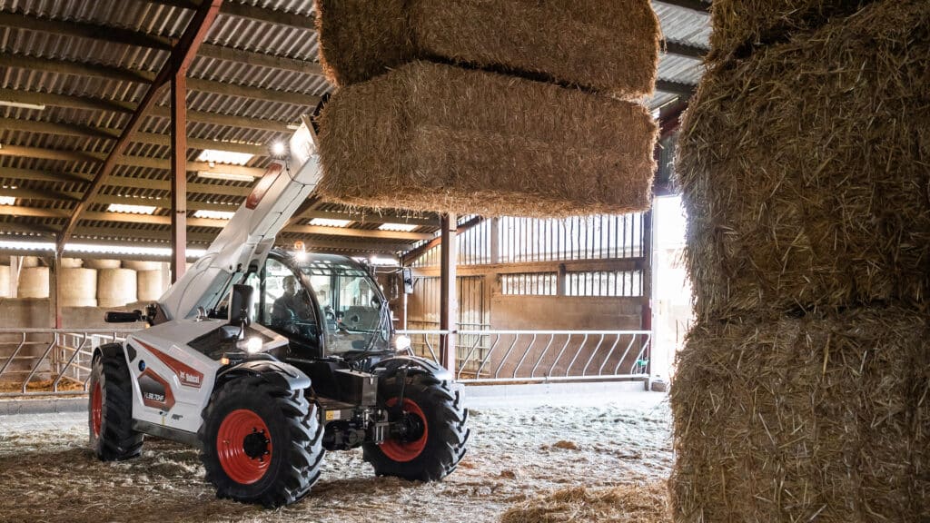 This image shows a Bobcat TL38.70 telehandler, a type of farm machinery used for heavy lifting and material handling on agricultural properties. The machine is lifting large bales of hay, showcasing its powerful lifting capacity and extendable boom. It is designed to handle a variety of farm tasks, such as stacking bales, moving feed, or transporting materials around the farm. The enclosed cabin provides comfort for the operator, making it ideal for long hours of use in agricultural settings.