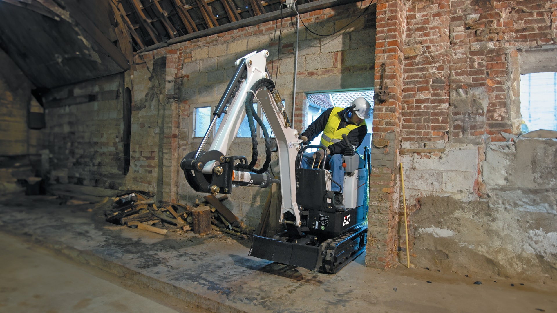 This image shows a Bobcat E10 mini excavator working inside a building, showcasing its compact size and versatility. The excavator is small enough to operate in confined indoor spaces, making it ideal for demolition, renovation, and construction tasks within tight or restricted areas. The operator is using the machine to perform precise digging, taking advantage of its zero tail swing design that allows for efficient manoeuvring without hitting walls. This mini excavator is perfect for interior work, where space and accessibility are often limited.