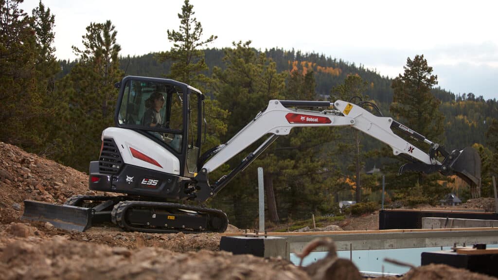 This image captures a Bobcat E35 mini excavator at a construction site in a forested, mountainous area. The operator is focused on using the machine's long arm attachment for precise digging and material handling. The compact design of the E35, combined with its powerful capabilities, makes it ideal for working in rugged outdoor environments where manoeuvrability and reliability are essential. This mini excavator is suited for tasks such as foundation work, trenching, and site preparation in remote or challenging terrains.