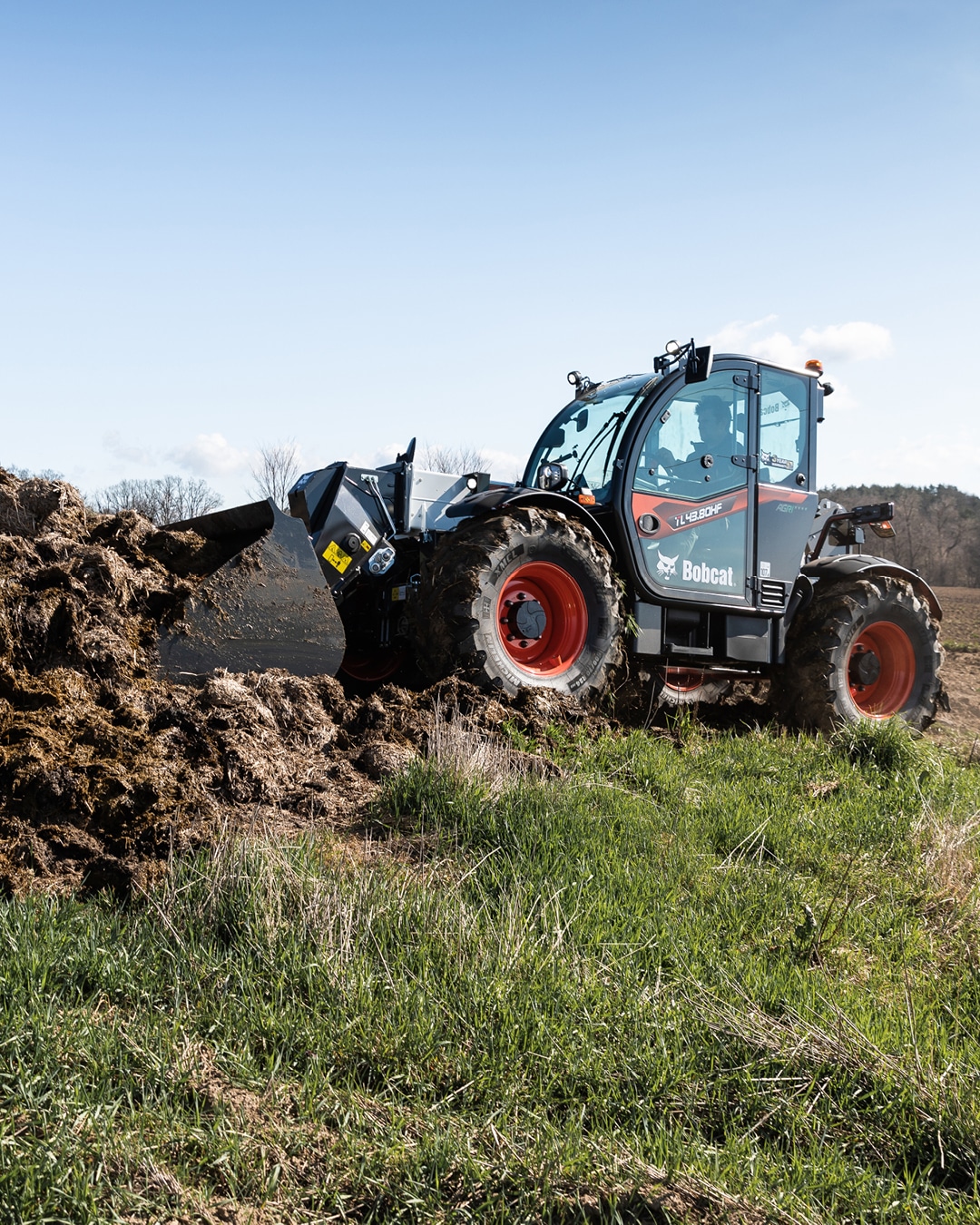 A Bobcat telehandler operating in an agricultural field, equipped with a bucket attachment, efficiently handling a pile of organic material. Its rugged build and bright red wheels stand out against the lush green grass and open sky, emphasising its reliability and versatility for farm-related tasks in outdoor environments.