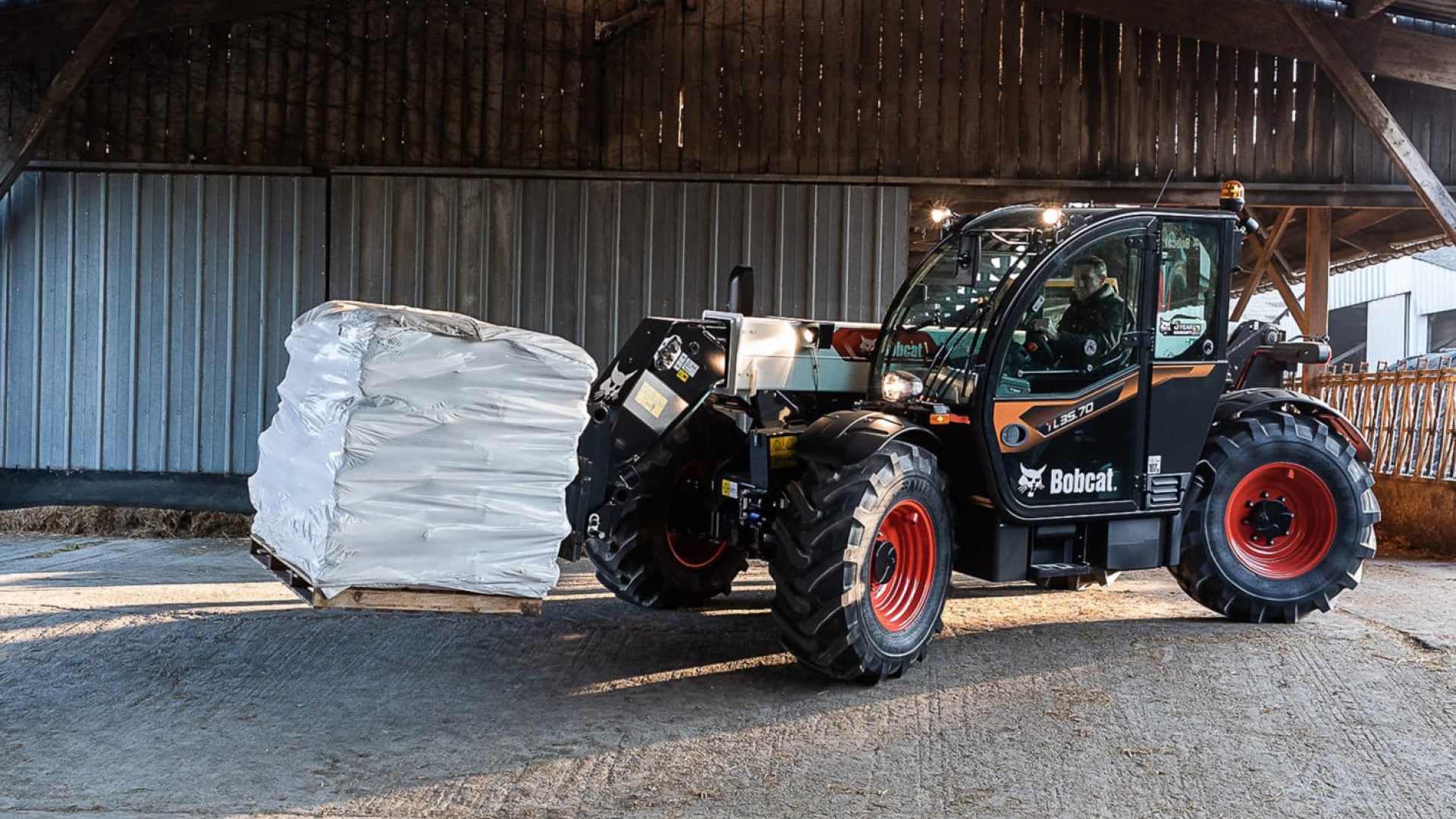 A Bobcat telehandler lifting a wrapped pallet of supplies inside an agricultural barn. The machine's sleek black design with bright red wheels stands out as it handles the heavy load with precision. The barn setting highlights the telehandler's practicality and efficiency for material handling tasks in farm environments.
