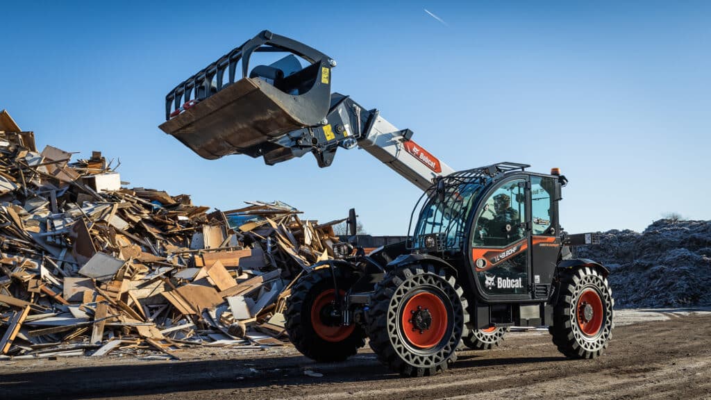 A Bobcat telehandler operating at a waste facility, equipped with a grapple bucket, efficiently lifting and managing a large pile of discarded materials and debris. The machine's rugged design and bright red wheels highlight its durability and versatility for heavy-duty tasks, with the clear blue sky providing a contrasting backdrop to the busy waste management environment.