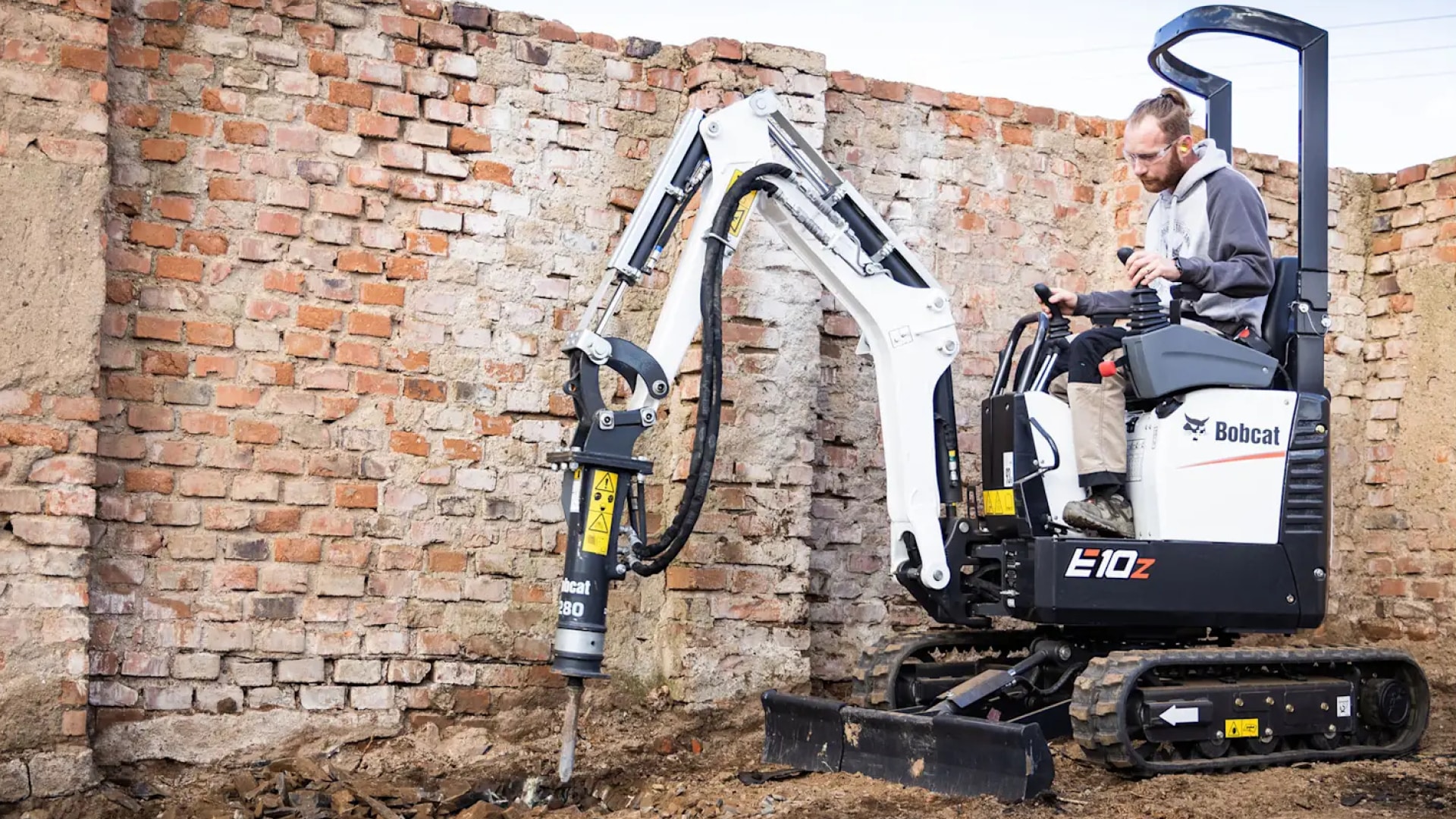 Operator using the Bobcat E10 Mini Excavator equipped with a hydraulic breaker attachment, demonstrating its power and efficiency for construction tasks against a brick wall background.
