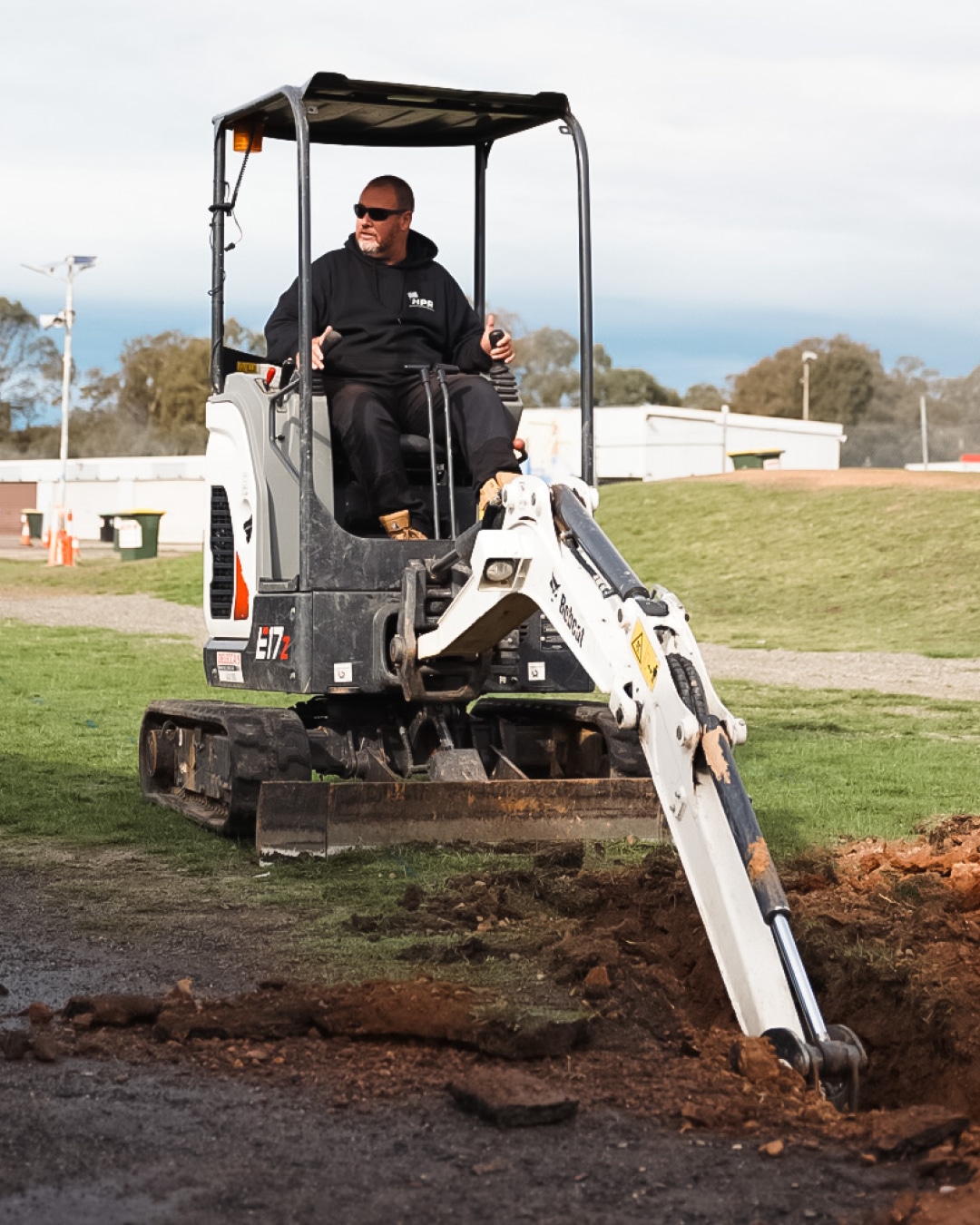 A man operating a Bobcat E17 mini excavator on a grassy field, excavating soil and creating a trench. The setting includes a background with buildings, trees, and construction elements, showcasing the machine's capability for small-scale excavation projects.