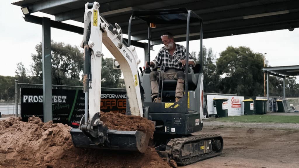 A man operating a Bobcat E17 mini excavator under a covered structure, scooping a large pile of dirt. The background features commercial banners and green bins, indicating an organised worksite. Trees and open space are visible in the distance.