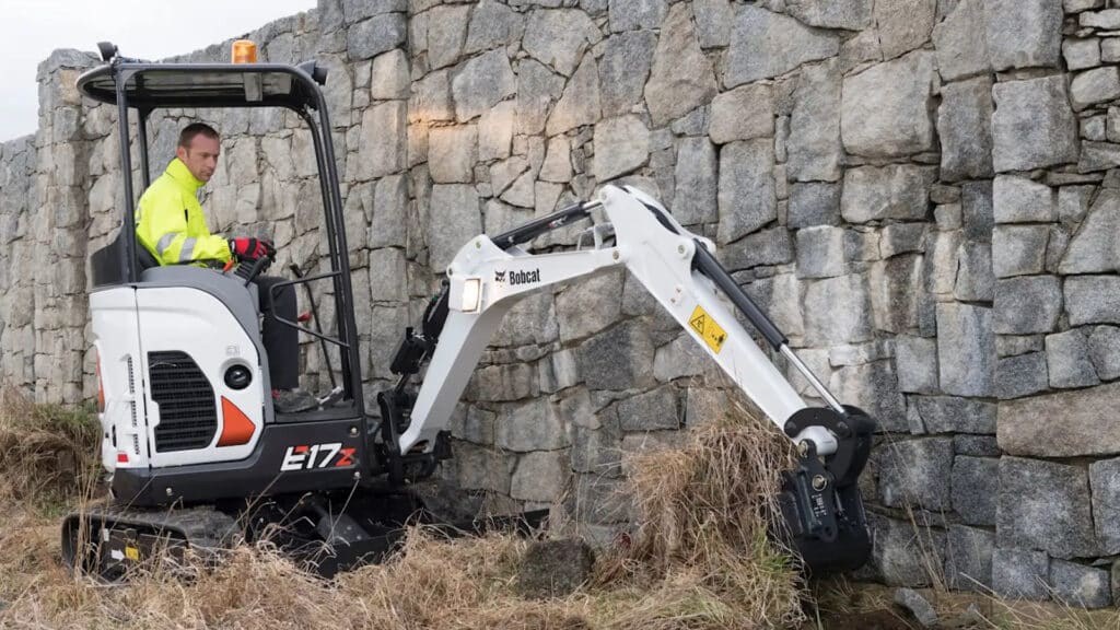 A worker in a high-visibility jacket operating a Bobcat E17 mini excavator near a stone wall, excavating overgrown grass and soil. The setting highlights the machine's precision and ability to work in confined spaces, with the rugged stone backdrop adding to the scene's industrial atmosphere.