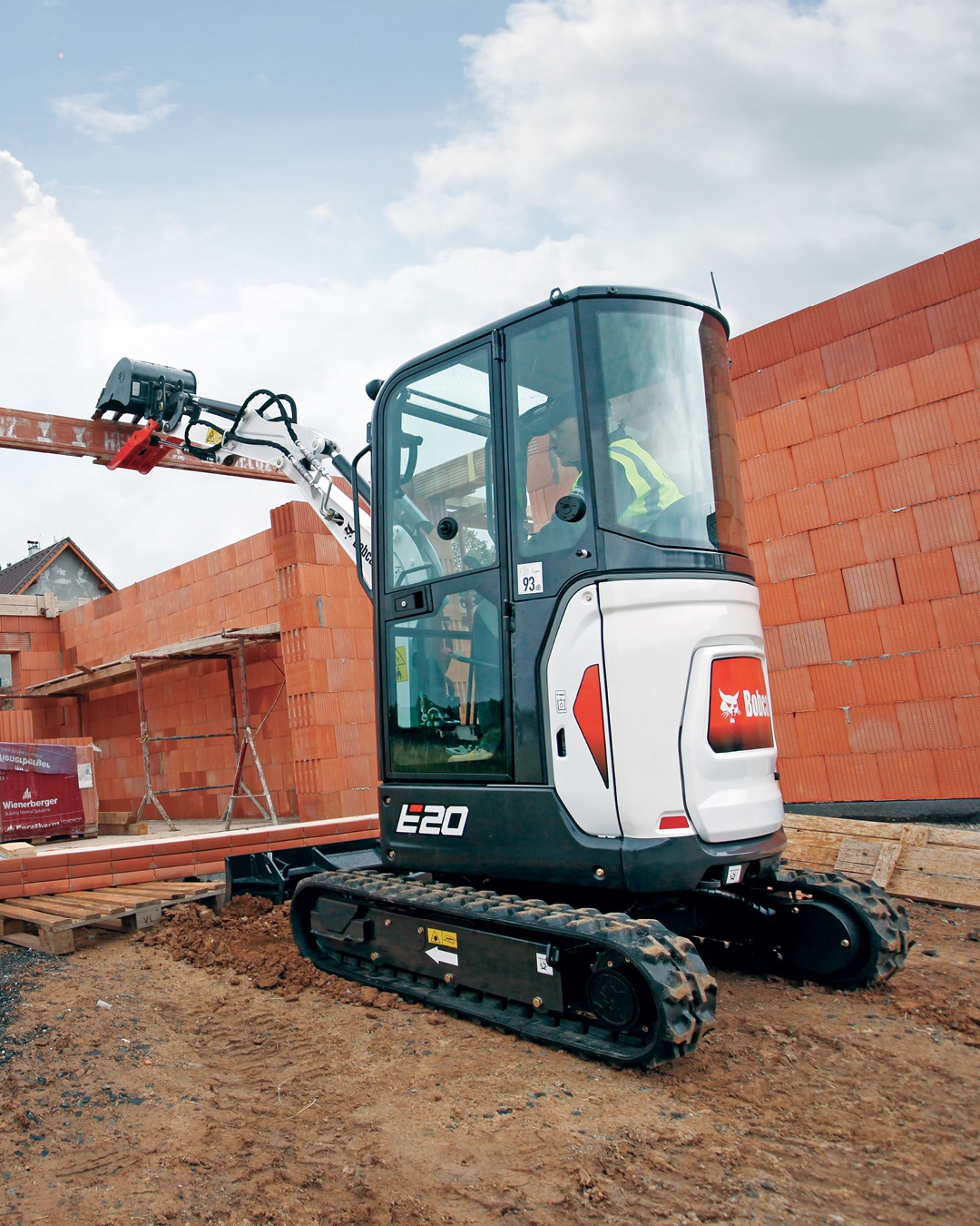 The image features a Bobcat E20 Mini Excavator at a construction site, showcasing its compact and agile design. The machine is equipped with an enclosed cab, an optional upgrade, and is being operated on a foundation of freshly prepared soil. Surrounded by brick walls and building materials, the excavator highlights its suitability for precise tasks in tight or urban spaces.