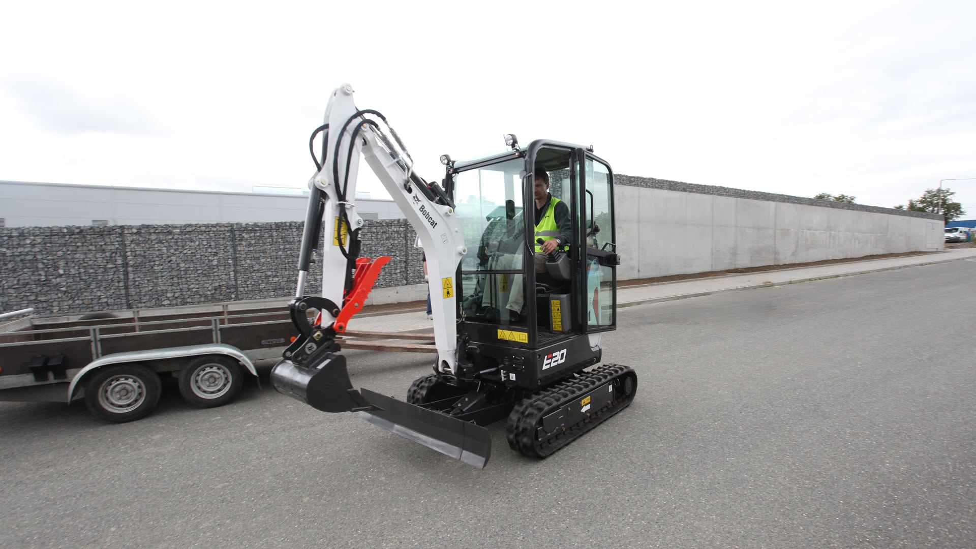 The image features a Bobcat E20 Mini Excavator on a paved road. The machine is operated by a person seated in an enclosed cab, demonstrating the optional cab upgrade. It is equipped with a digging bucket and a front blade, highlighting its versatility for various applications. The excavator’s compact design makes it ideal for urban or confined job sites.