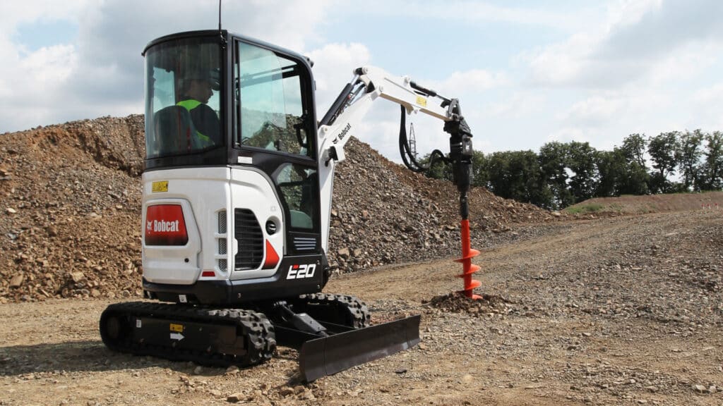 The image showcases a Bobcat E20 Mini Excavator equipped with an auger attachment, actively drilling into the ground on a construction site. The machine is operated by a person seated in an enclosed cab, illustrating the optional cab feature. Its compact design and versatility are highlighted, making it ideal for earthmoving tasks in confined or open spaces.