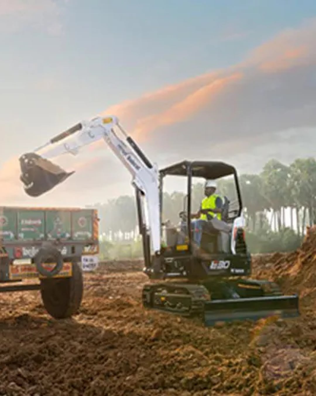 The image features a Bobcat E30 Mini Excavator working on a construction site during sunset, with the operator loading soil into a nearby truck. The machine is shown in action, equipped with a digging bucket and an open canopy, highlighting its efficiency and versatility for earthmoving tasks in various environments. The scenic backdrop enhances the depiction of a productive work setting.