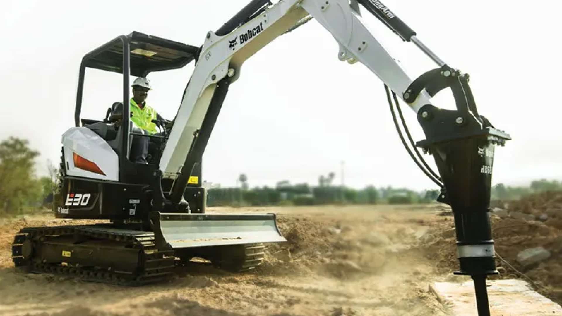The image shows a Bobcat E30 Mini Excavator equipped with a hydraulic breaker attachment, demonstrating its versatility for demolition and breaking tasks. The operator is seated in an open canopy, efficiently controlling the machine on a flat construction site. This highlights the E30's adaptability and performance in handling demanding applications with precision.
