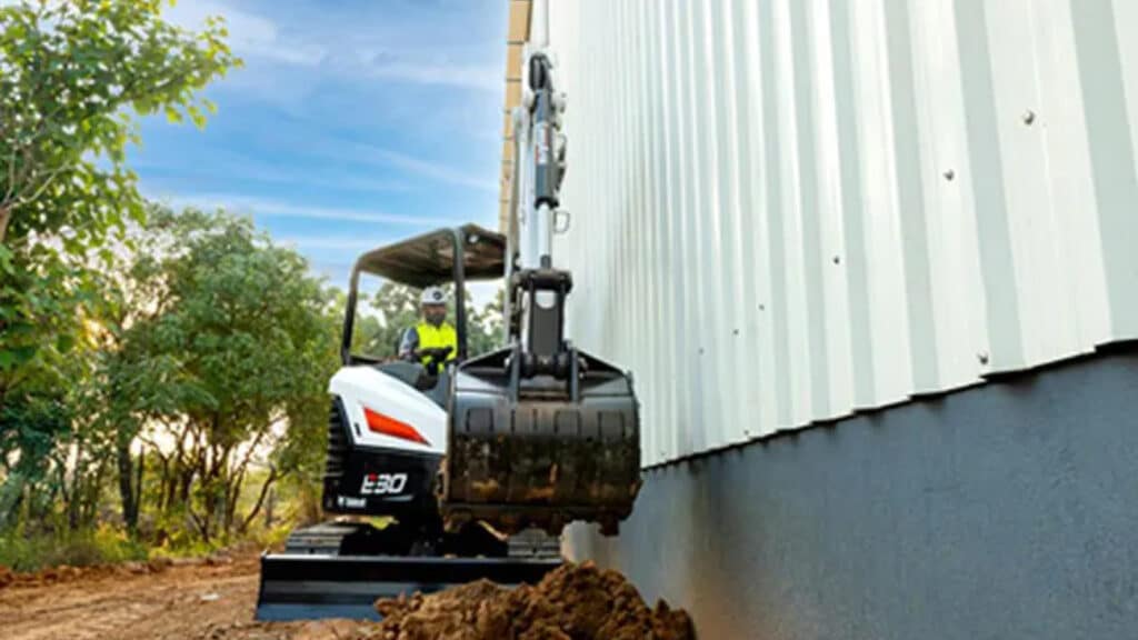The image showcases a Bobcat E30 Mini Excavator working efficiently in a tight space along the side of a building. The operator, seated in an open canopy, skilfully manoeuvres the machine while utilising the digging bucket for precise excavation. This highlights the E30's compact design and suitability for operations in confined areas without compromising performance.