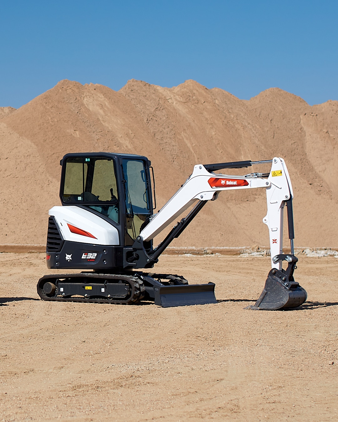 The image features a Bobcat E32 Mini Excavator positioned on a sandy worksite, with large mounds of dirt in the background. The machine is shown stationary, equipped with an enclosed cab for operator comfort and a durable digging bucket. The scene highlights the E32's suitability for earthmoving and excavation projects in open and arid environments.