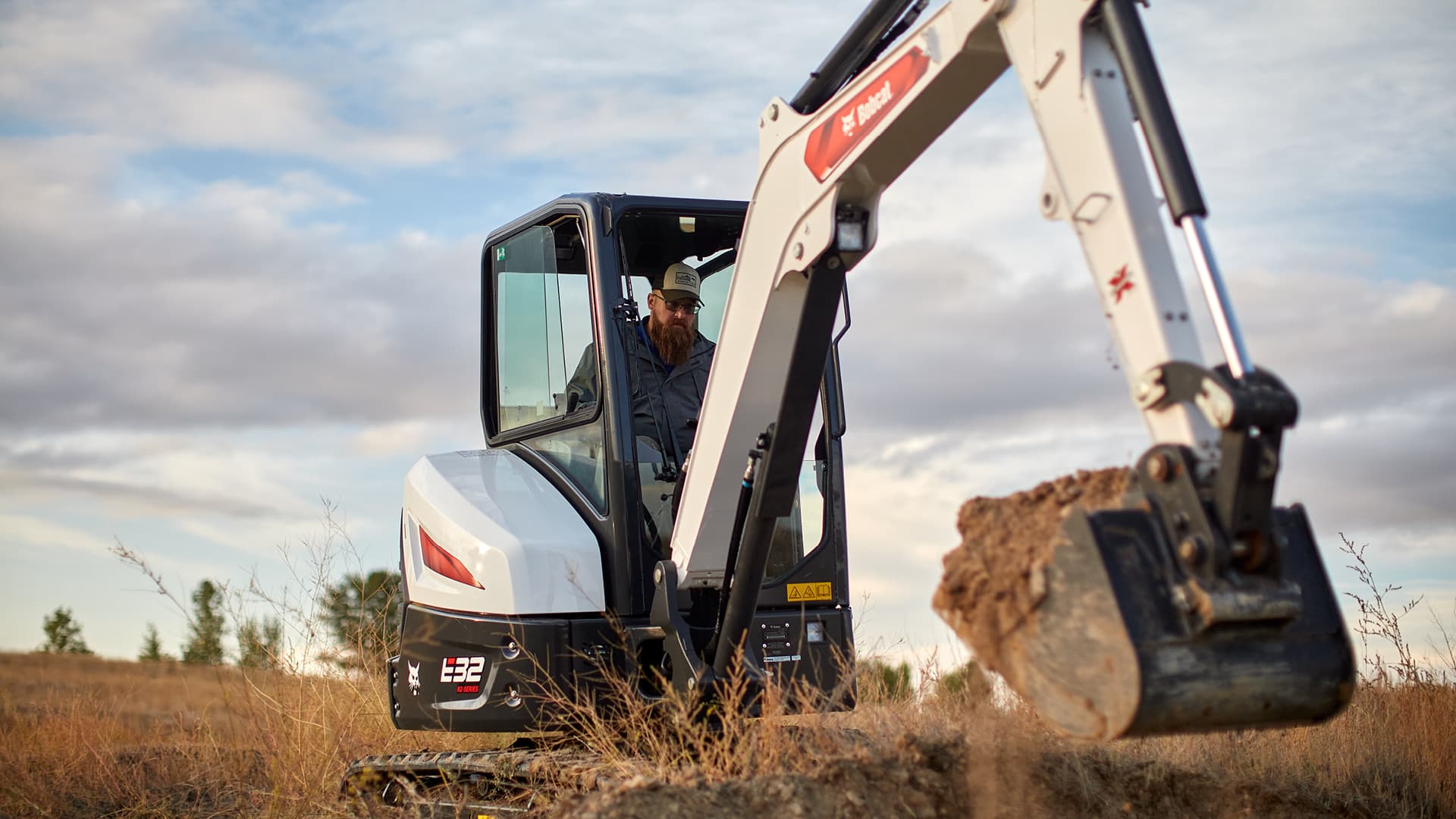 The image showcases a Bobcat E32 Mini Excavator at work on a rural site. The operator, seated in an enclosed cab, is concentrating as the machine's digging bucket handles a load of soil. The scenic backdrop of an open field and cloudy sky underscores the E32's ability to perform efficiently in outdoor and off-road environments, making it an ideal choice for versatile excavation tasks.