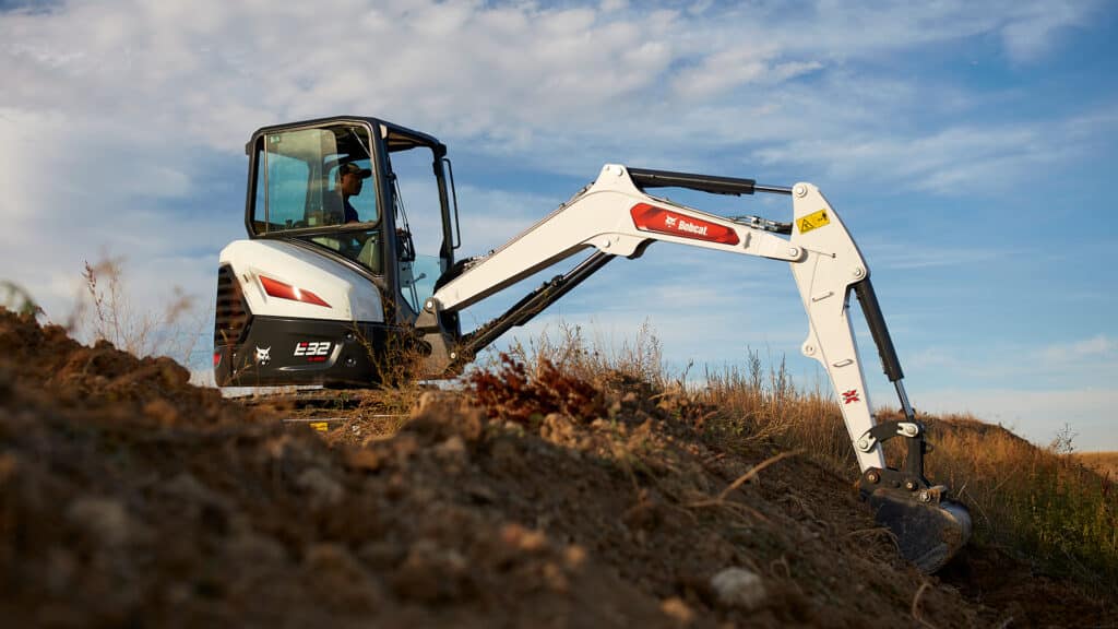 The image shows a Bobcat E32 Mini Excavator actively digging on a rugged hillside. The operator, seated in an enclosed cab, is efficiently controlling the arm and bucket, demonstrating the machine's precision and stability. The clear blue sky and natural terrain emphasise the E32's capability to handle challenging outdoor excavation tasks with ease and reliability.