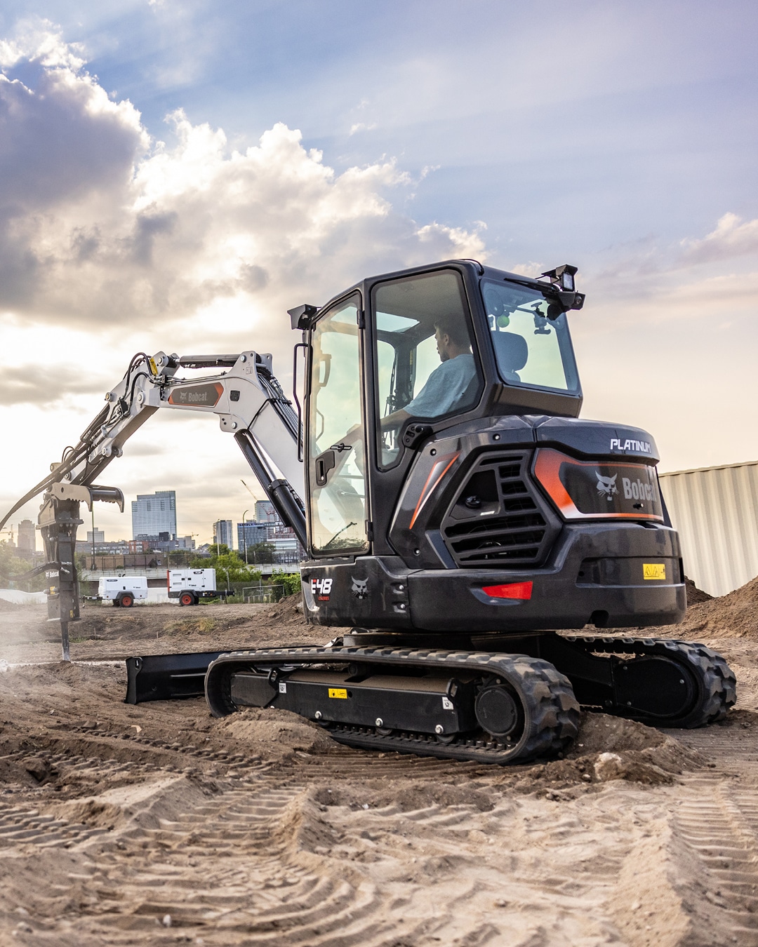 The image showcases a Bobcat E48 Mini Excavator Platinum Edition operating at a construction site. The machine is equipped with an enclosed cab for enhanced operator comfort, while its sleek design and advanced features are on display. The urban skyline and dramatic clouds in the background highlight the E48's capability to perform efficiently in modern and demanding construction environments.