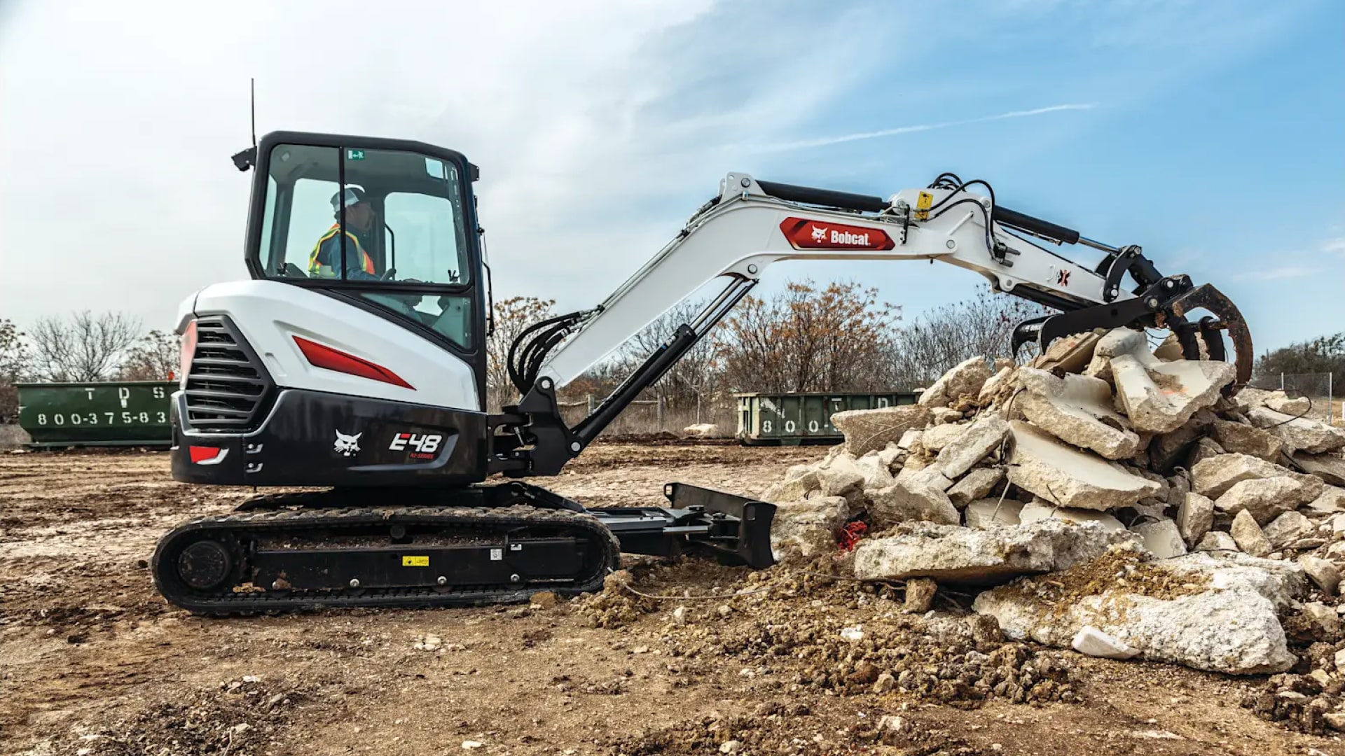 The image showcases a Bobcat E48 Mini Excavator in action at a construction site. The machine is equipped with a grapple attachment, efficiently handling a pile of concrete debris. The operator, seated in an enclosed cab, demonstrates the excavator's precision and power in managing heavy-duty material handling tasks. The setting emphasises the E48's versatility and reliability for demolition and site preparation projects.