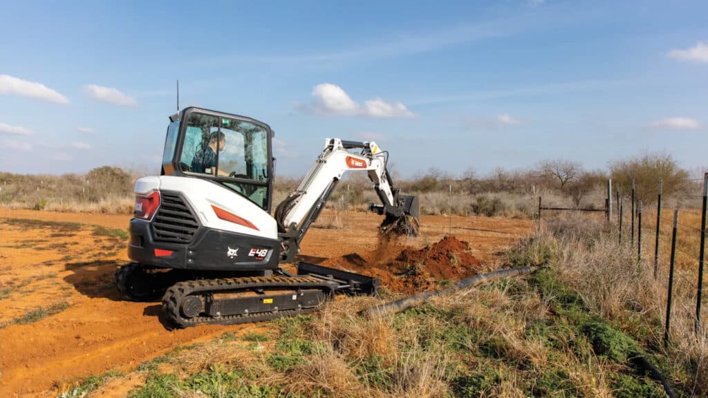 The image features a Bobcat E48 Mini Excavator actively digging in a rural area. The operator, seated in an enclosed cab, is efficiently managing the machine as it moves soil. The open field and clear sky emphasise the E48's adaptability and reliability for agricultural and land preparation tasks in outdoor environments.