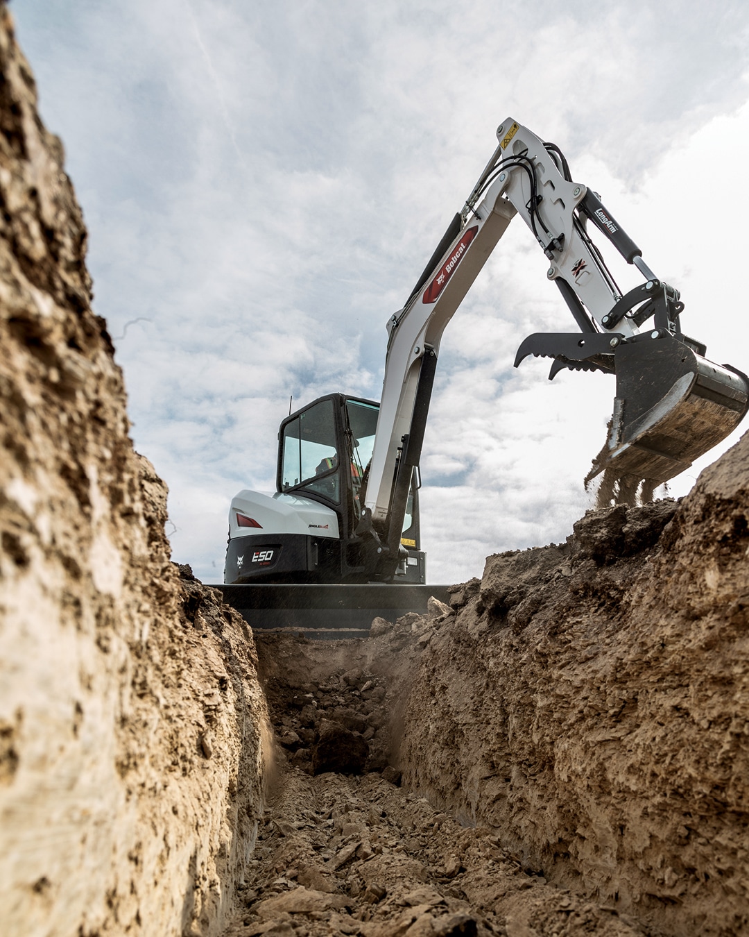 The image features a Bobcat E50 Mini Excavator working in a narrow trench, expertly excavating soil with its digging bucket. The operator, seated in an enclosed cab, is demonstrating the machine's precision and efficiency in tight spaces. The rugged terrain and cloudy sky emphasise the E50's capability to handle challenging excavation tasks with ease and reliability.
