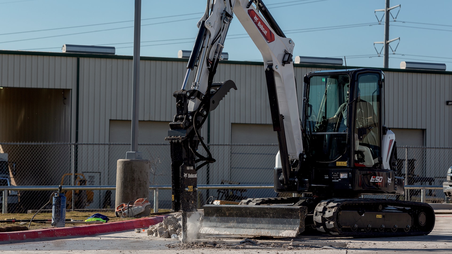 A Bobcat E50 Mini Excavator equipped with a hydraulic breaker attachment, actively working on a concrete surface. The operator, seated in an enclosed cab, demonstrates the excavator's precision and power, effectively breaking through tough material. The industrial setting highlights the E50's suitability for heavy-duty demolition and site preparation tasks.