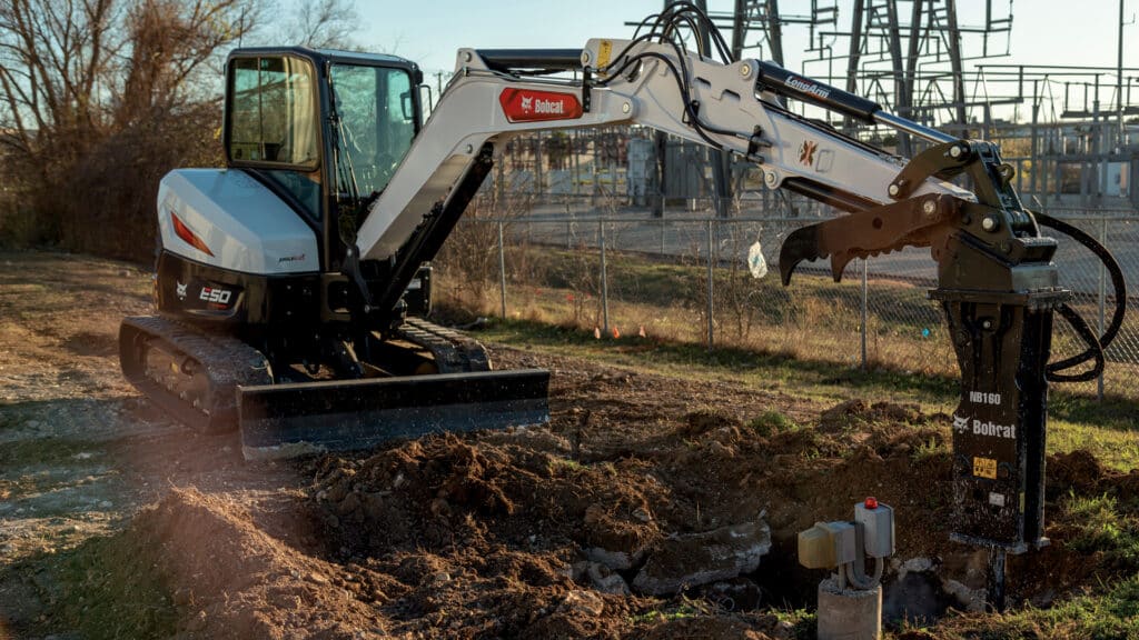 The image showcases a Bobcat E50 Mini Excavator equipped with a hydraulic breaker attachment, performing precise demolition work on a rural site. The machine, operated from an enclosed cab, is breaking through concrete with efficiency. The industrial background and natural surroundings emphasise the E50's versatility and ability to handle demanding tasks in diverse environments.