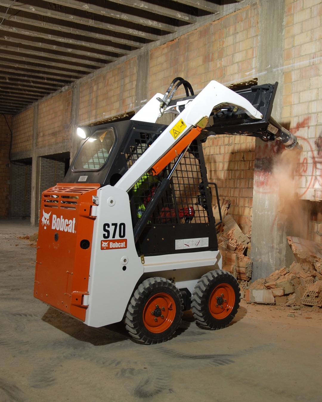 A Bobcat S70 skid steer loader equipped with a hydraulic breaker attachment, expertly demolishing a brick wall inside a construction site with precision and power.