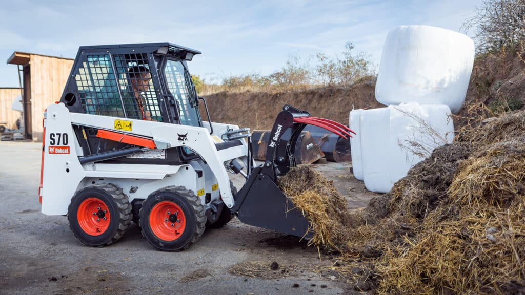 A Bobcat S70 skid steer loader handling straw and organic material with a grapple bucket, working efficiently in a rural setting near stacked silage bales.