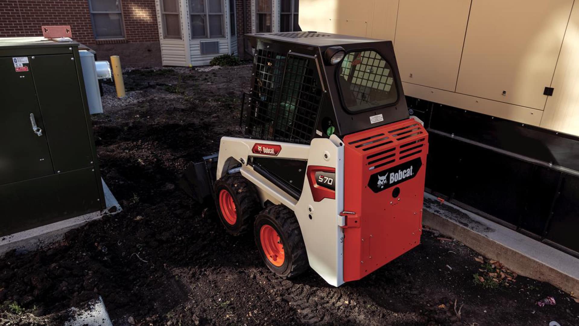 A Bobcat S70 skid steer loader working in a compact urban space, efficiently handling soil and landscaping tasks in a tight area near buildings and utility equipment.