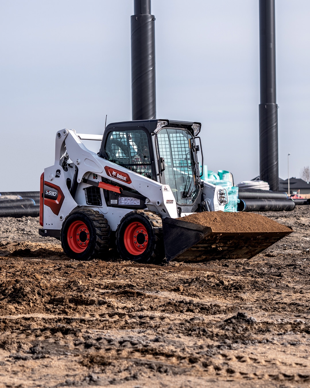 A Bobcat Skid Steer Loader operating on a construction site, equipped with a bucket attachment and moving a load of soil, showcasing its power and efficiency in earthmoving tasks under clear skies.