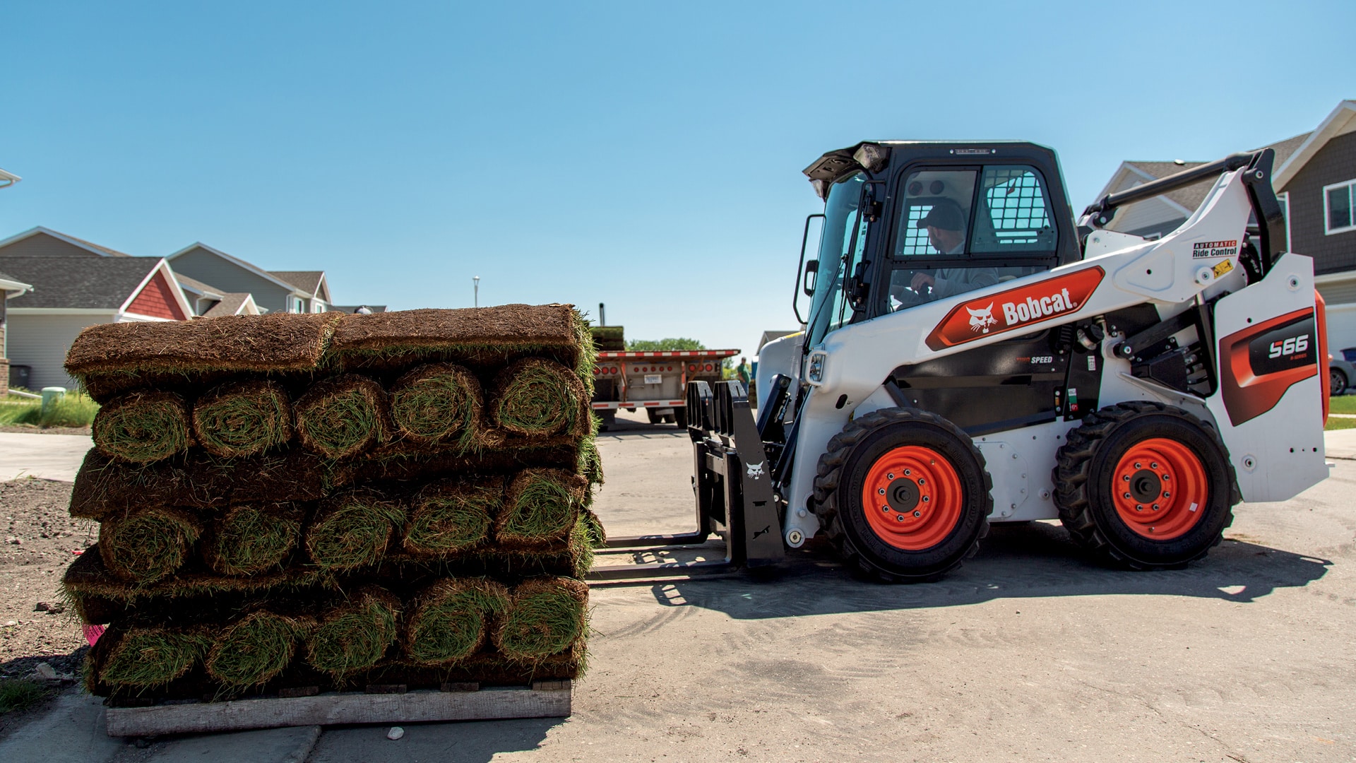 A Bobcat S66 skid-steer loader equipped with a pallet fork attachment is shown lifting a pallet of rolled turf. The loader is positioned in a residential area, highlighting its versatility for landscaping and construction tasks. The machine's compact design and precise control make it ideal for manoeuvring in tight spaces.