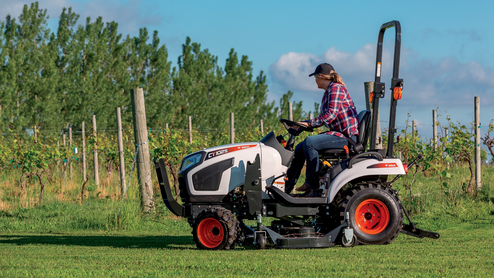 A Bobcat CT1025 compact tractor is featured in a lush, green vineyard, operated by a person in casual workwear. The image captures the tractor's versatility and ease of use, making it an ideal choice for small acreage owners and hobby farmers. The vibrant backdrop emphasises the connection between the tractor and its agricultural setting.