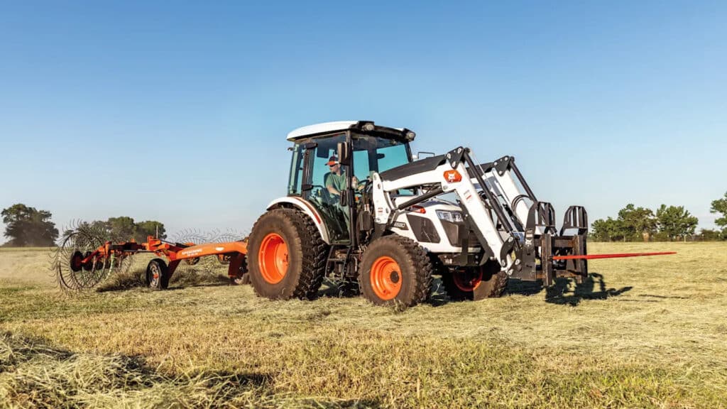 A Bobcat utility tractor is shown working in an open field, paired with a hay rake, efficiently preparing hay under a clear blue sky. The tractor's front loader is equipped with a hay spear, showcasing its versatility for farm tasks. The setting highlights its power, durability, and suitability for agricultural operations.