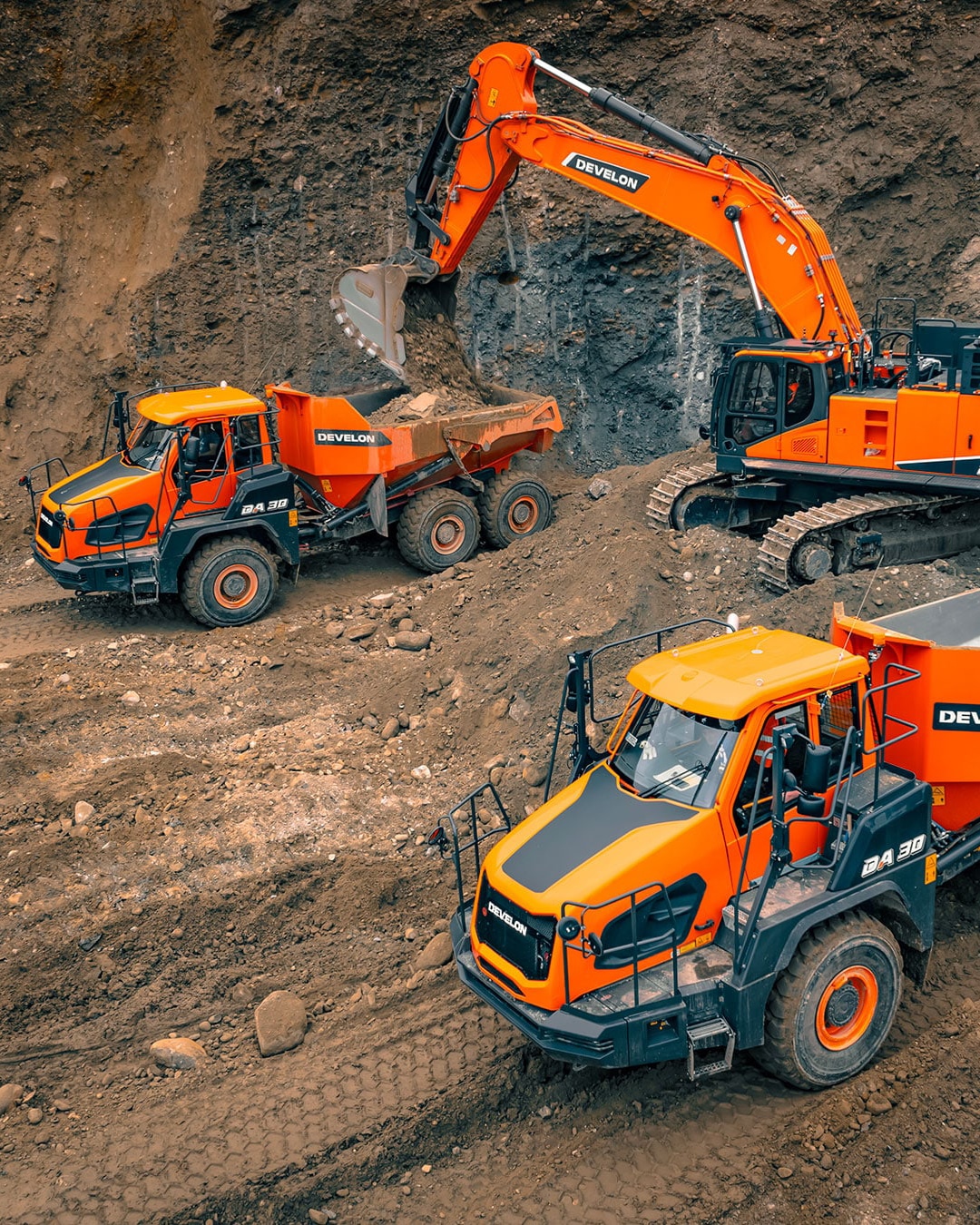 A construction site showcasing multiple Develon articulated dump trucks in action, with one being loaded by a Develon large excavator. The scene highlights their rugged design, large tyres, and efficient collaboration for moving heavy loads. The earthy background emphasises their capability to perform in tough and demanding environments.