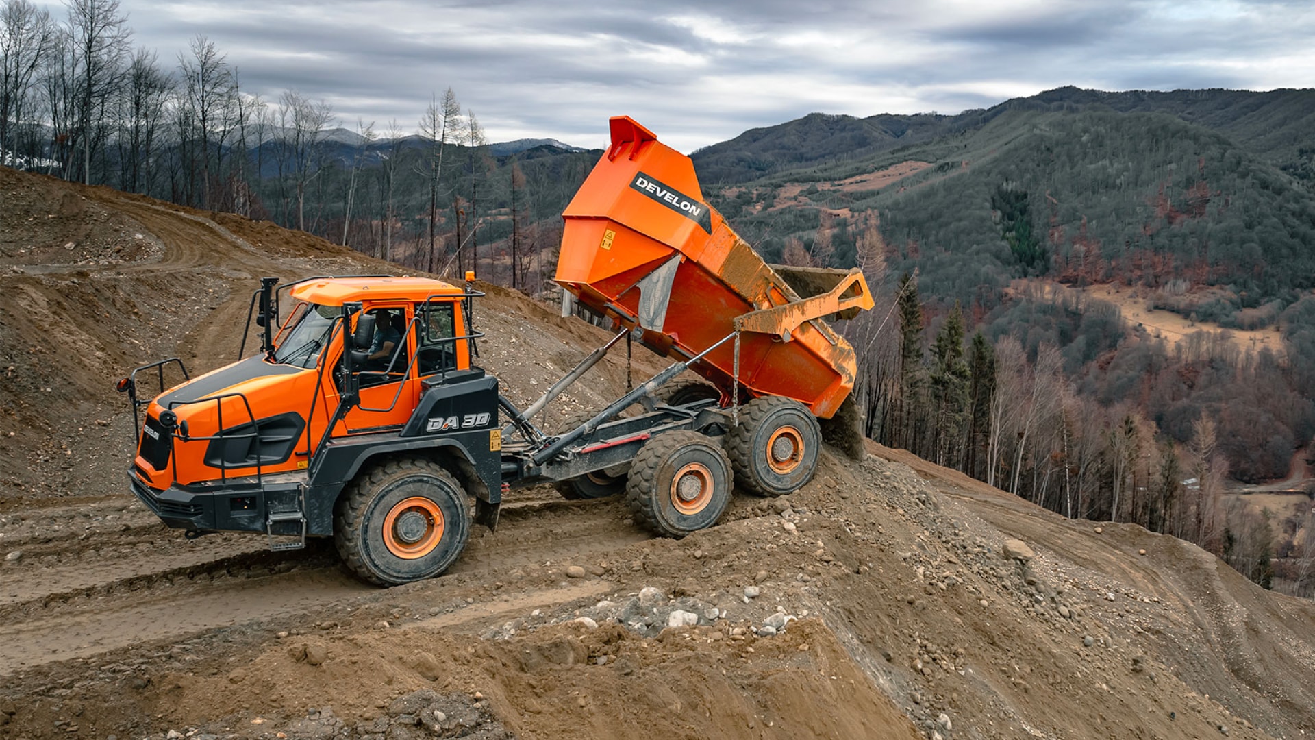 A Develon articulated dump truck unloading a full load of dirt on a rugged, muddy construction site. The truck's bright orange body and large tires stand out against a backdrop of rolling hills, dense forests, and overcast skies, emphasising its strength and versatility in challenging terrains.