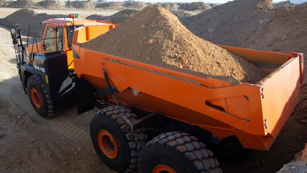 A close-up view of a Develon articulated dump truck fully loaded with a large pile of dirt. The bright orange dump bed contrasts against the rugged terrain of a construction site, highlighting the truck's heavy-duty capabilities. The large, durable tyres and sturdy frame emphasise its reliability for hauling in demanding environments.
