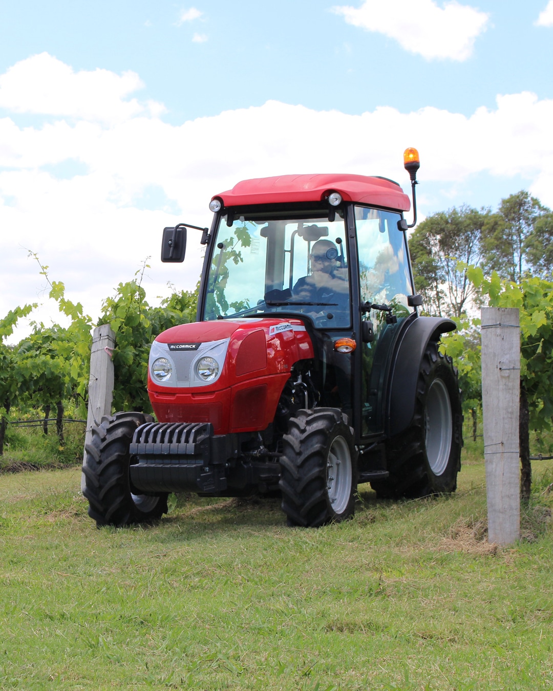 A McCormick tractor is displayed navigating through a lush vineyard, with its vibrant red finish standing out against the green vines and grass. The tractor's compact design and agility make it ideal for precision work in confined agricultural spaces, highlighting its efficiency and suitability for vineyard tasks.