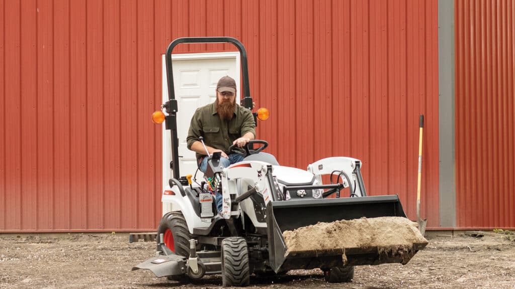A person operates a Bobcat CT1025 Compact Tractor carrying a bucket full of dirt in front of a red metal building, highlighting its power and versatility for small property tasks.