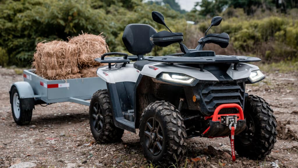 A Segway Farm Quad Bike with rugged tyres and front winch tows a small trailer loaded with hay bales across a dirt track, highlighting its power and utility for rural work.