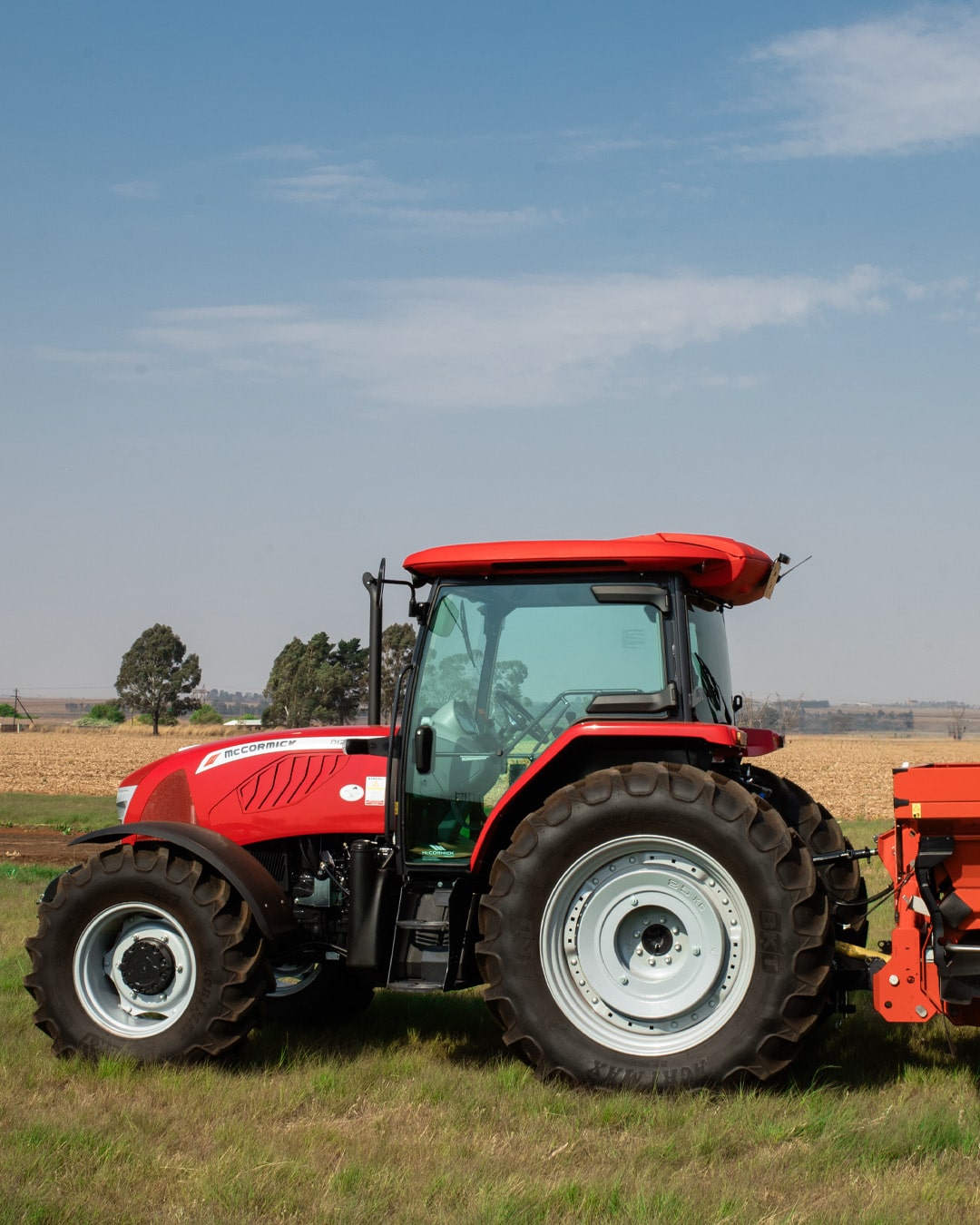 McCormick D-MAX Farm Tractor side view operating in a grassy field, attached to a rear-mounted implement, with open farmland and trees in the background under a clear sky.
