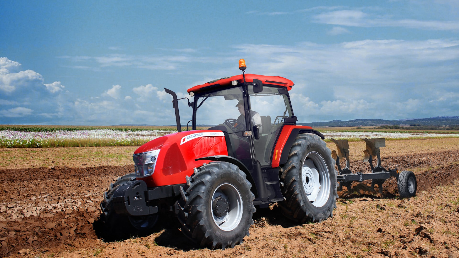 McCormick D-MAX Farm Tractor working in a ploughed field with a rear-mounted plough, surrounded by flowering crops and distant hills under a partly cloudy sky.