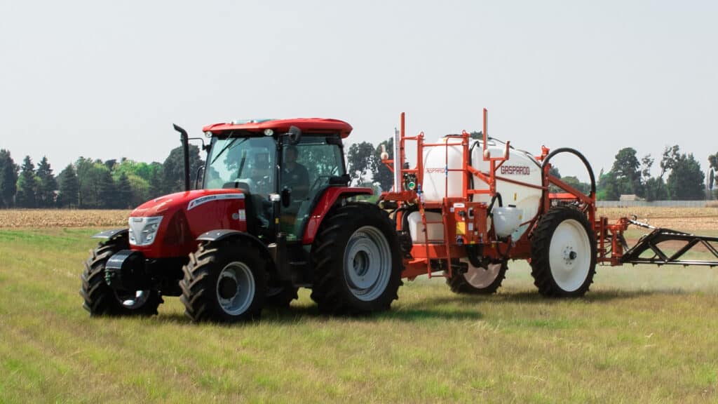 McCormick D-MAX Farm Tractor towing a Gaspardo sprayer through a grassy field with trees and farmland in the background.