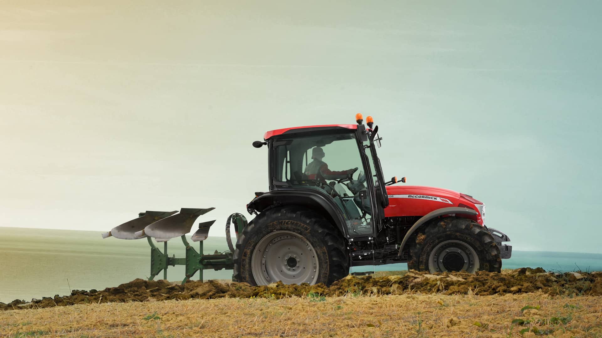 McCormick X5 Farm Tractor pulling a plough across a tilled field near the coastline at sunrise, showcasing its strength and adaptability on sloped terrain.
