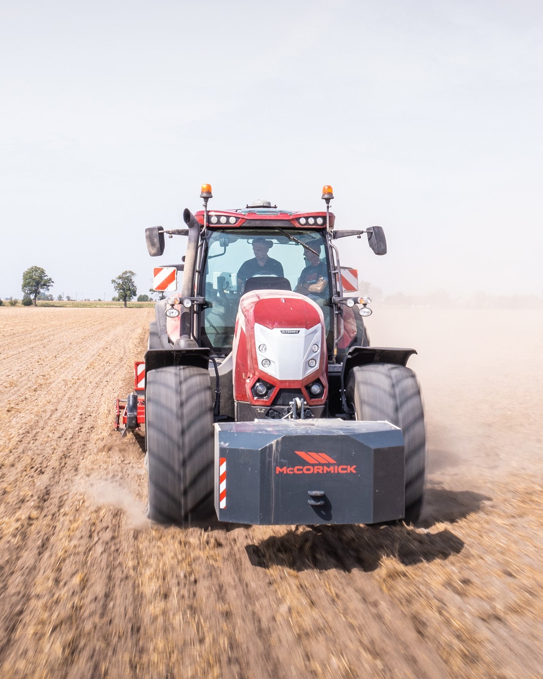 McCormick X8 Farm Tractor driving directly toward the camera across a wide, freshly cultivated paddock, kicking up dust while towing a red implement, with open farmland and scattered trees in the background.