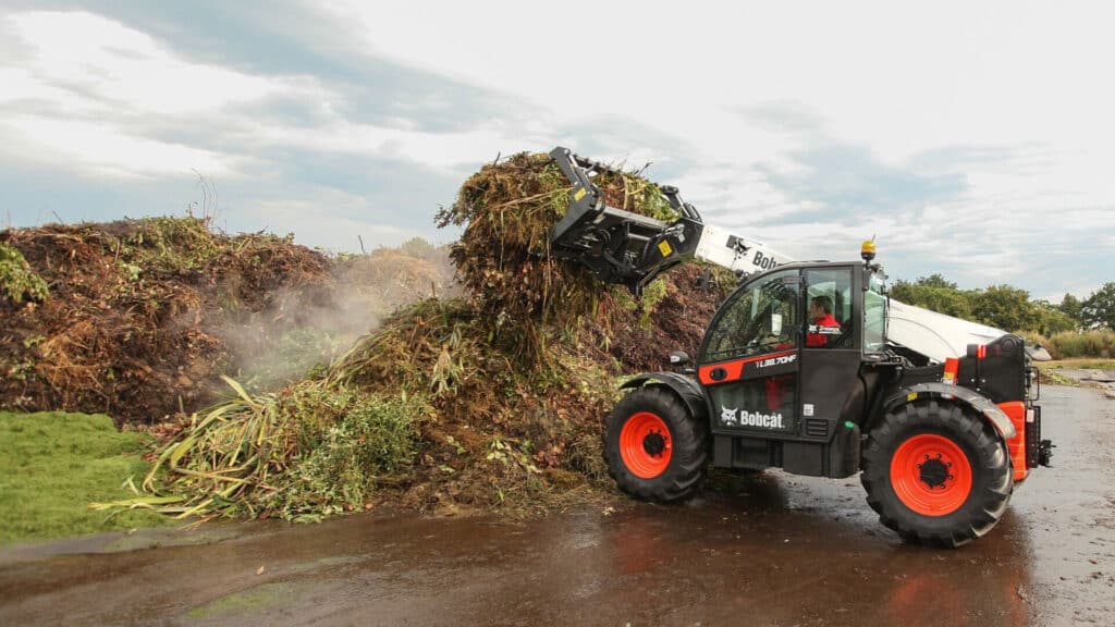 A Bobcat Agricultural Telehandler in action, lifting a steaming pile of organic vegetation with a grapple bucket on a composting or green waste site.