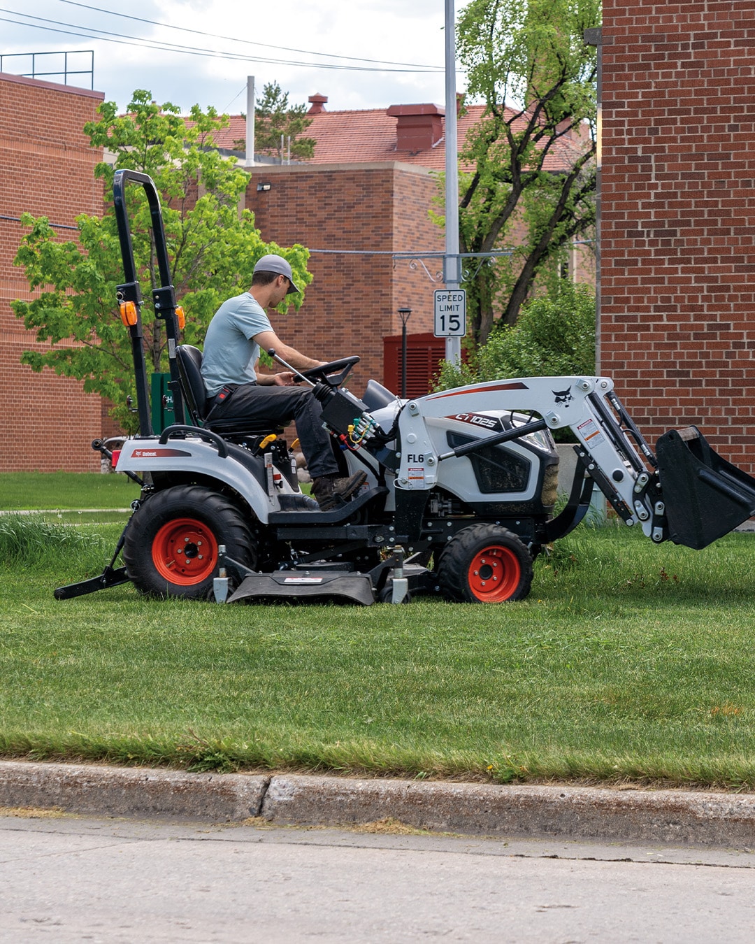 A person operates a Bobcat CT1025 Compact Tractor with a front-end loader and mid-mount mower on a grassy area near brick buildings, ideal for urban property maintenance and landscaping tasks.