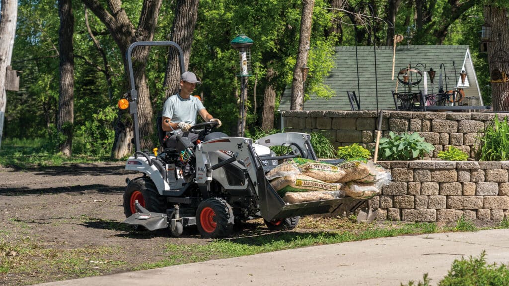 A person operates a Bobcat CT1025 Compact Tractor carrying bags of wood mulch in the front-end loader, working near a raised garden bed in a well-kept yard surrounded by trees.