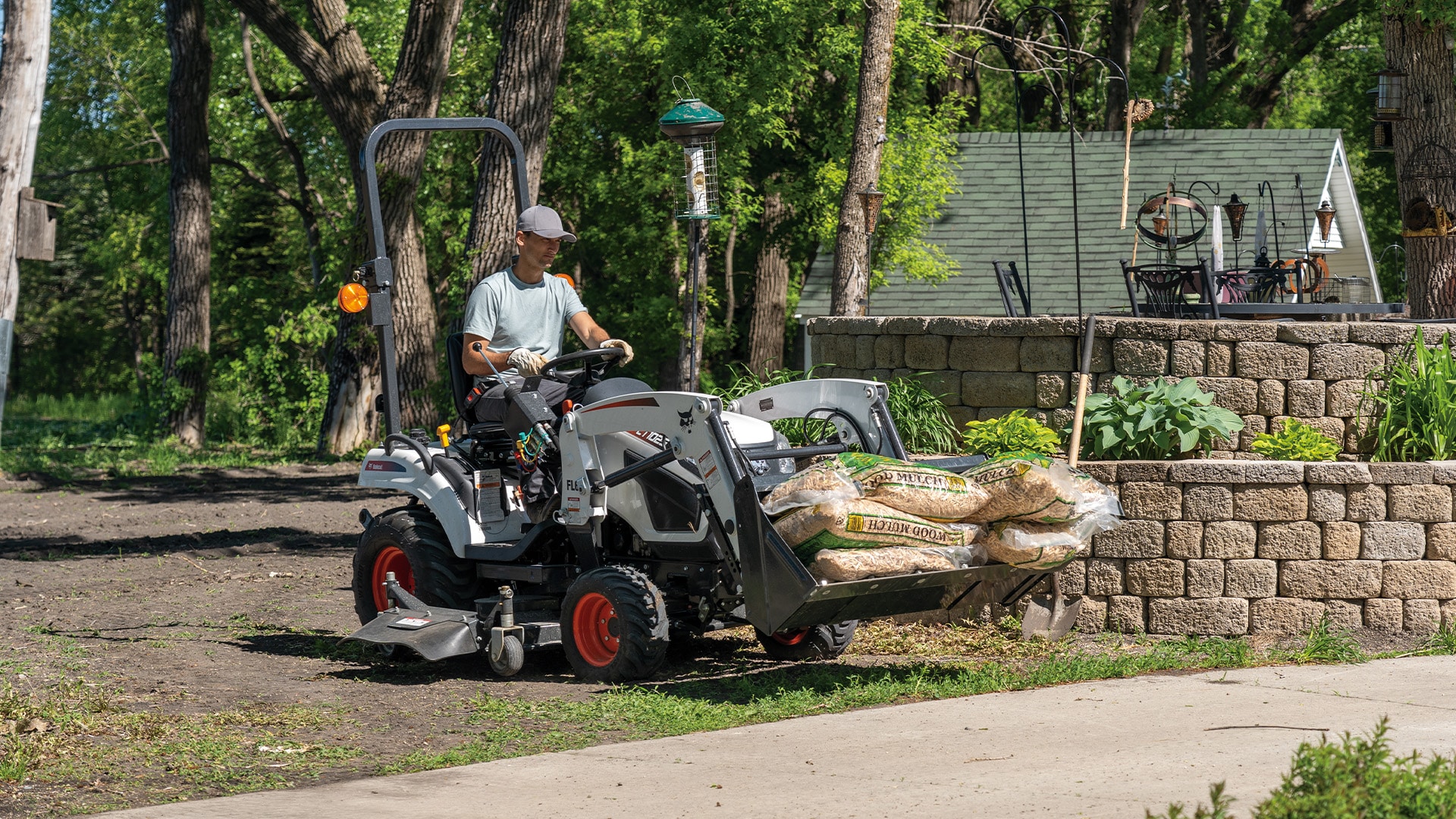 A person operates a Bobcat CT1025 Compact Tractor carrying bags of wood mulch in the front-end loader, working near a raised garden bed in a well-kept yard surrounded by trees.